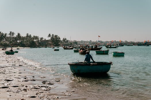 Tranquil scene of a fisherman on a round boat in Mui Ne Beach, Vietnam, with palm trees and boats under a clear sky.