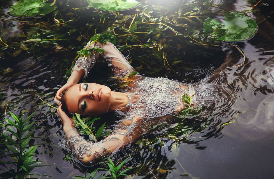 A woman in a shimmering dress peacefully floats in a lush pond surrounded by water plants.
