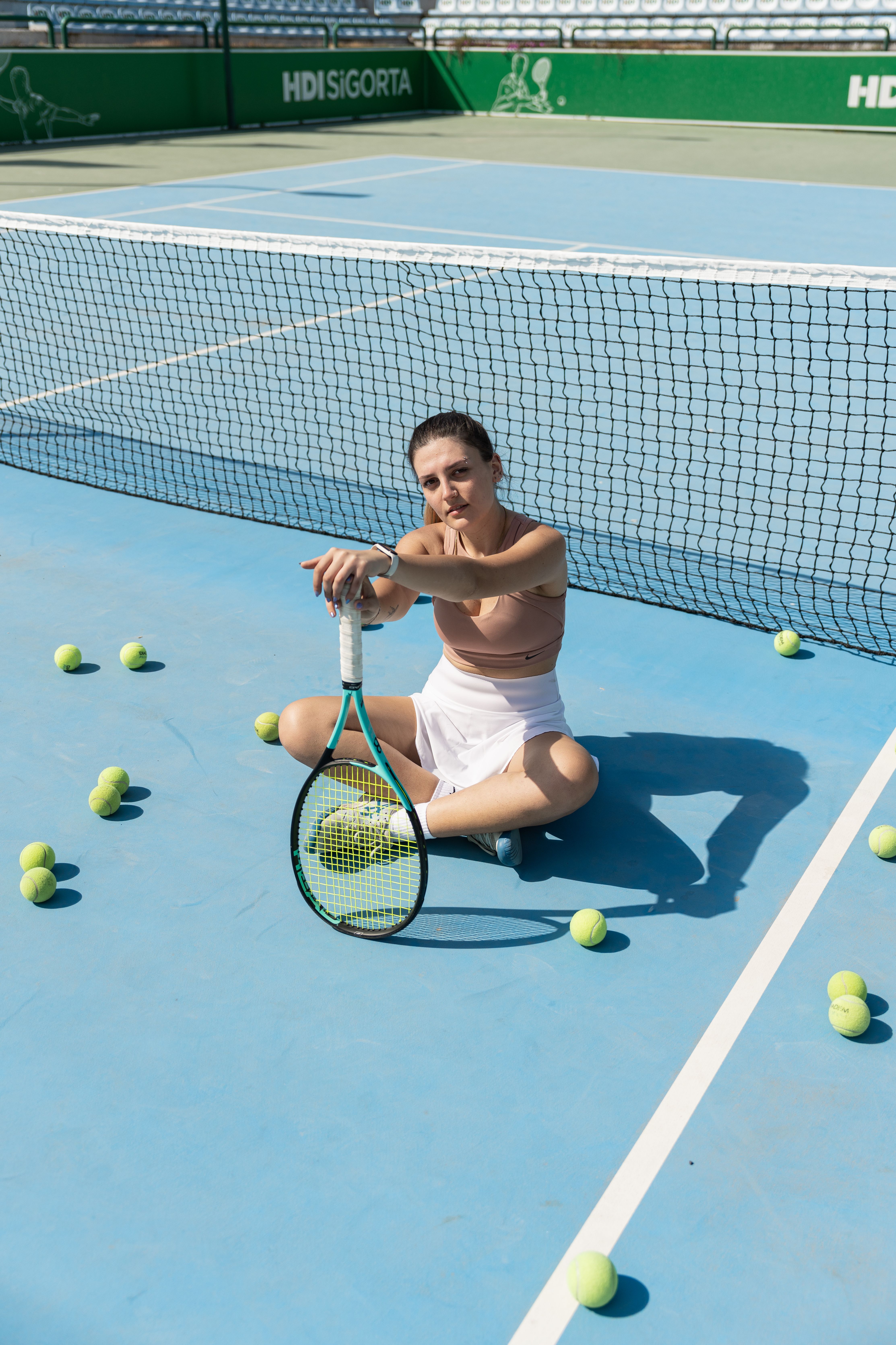 Woman in sportswear sitting on tennis court surrounded by balls after a match.
