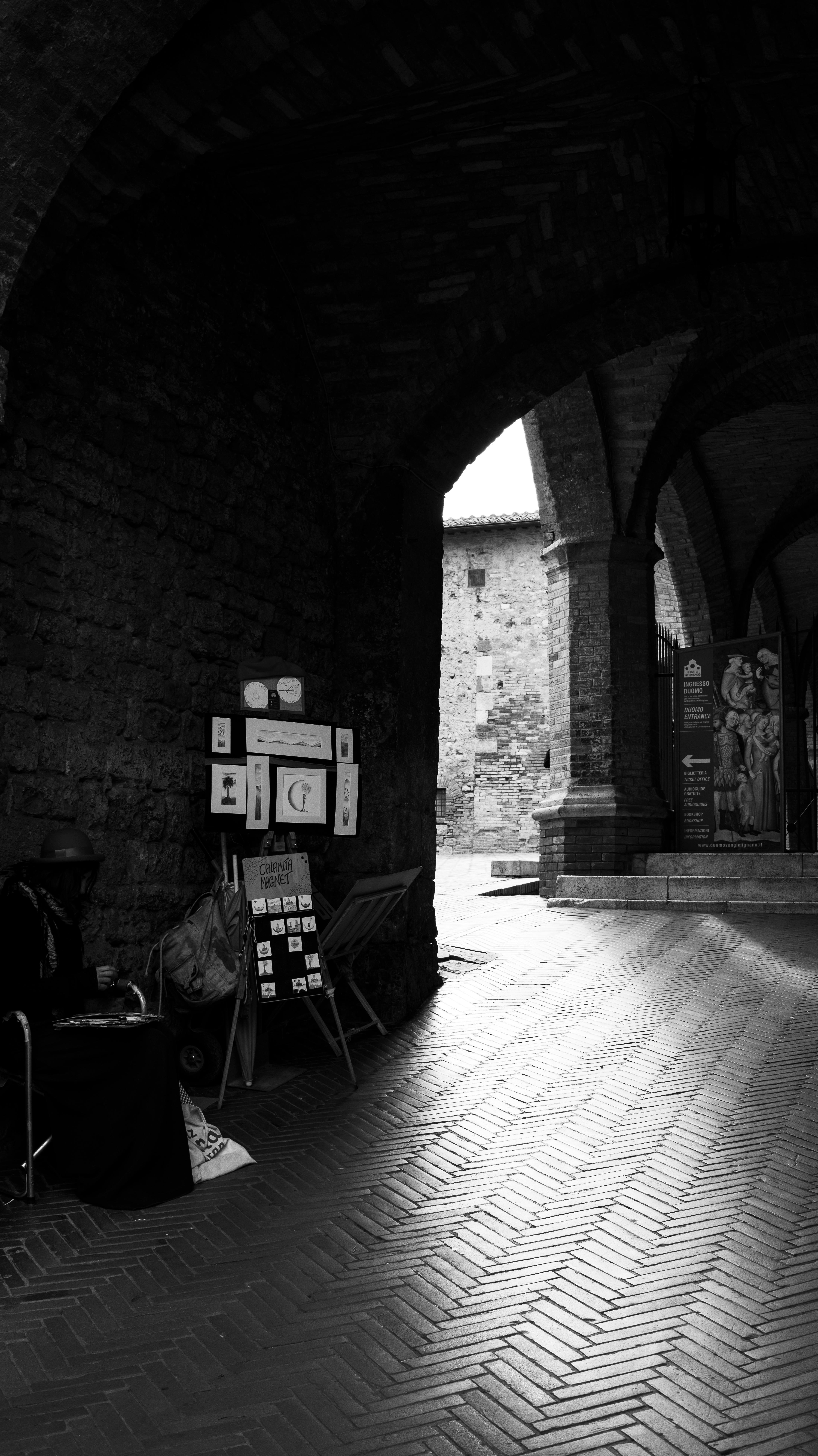A black and white view of a historic stone archway featuring an art display and cobbled flooring.