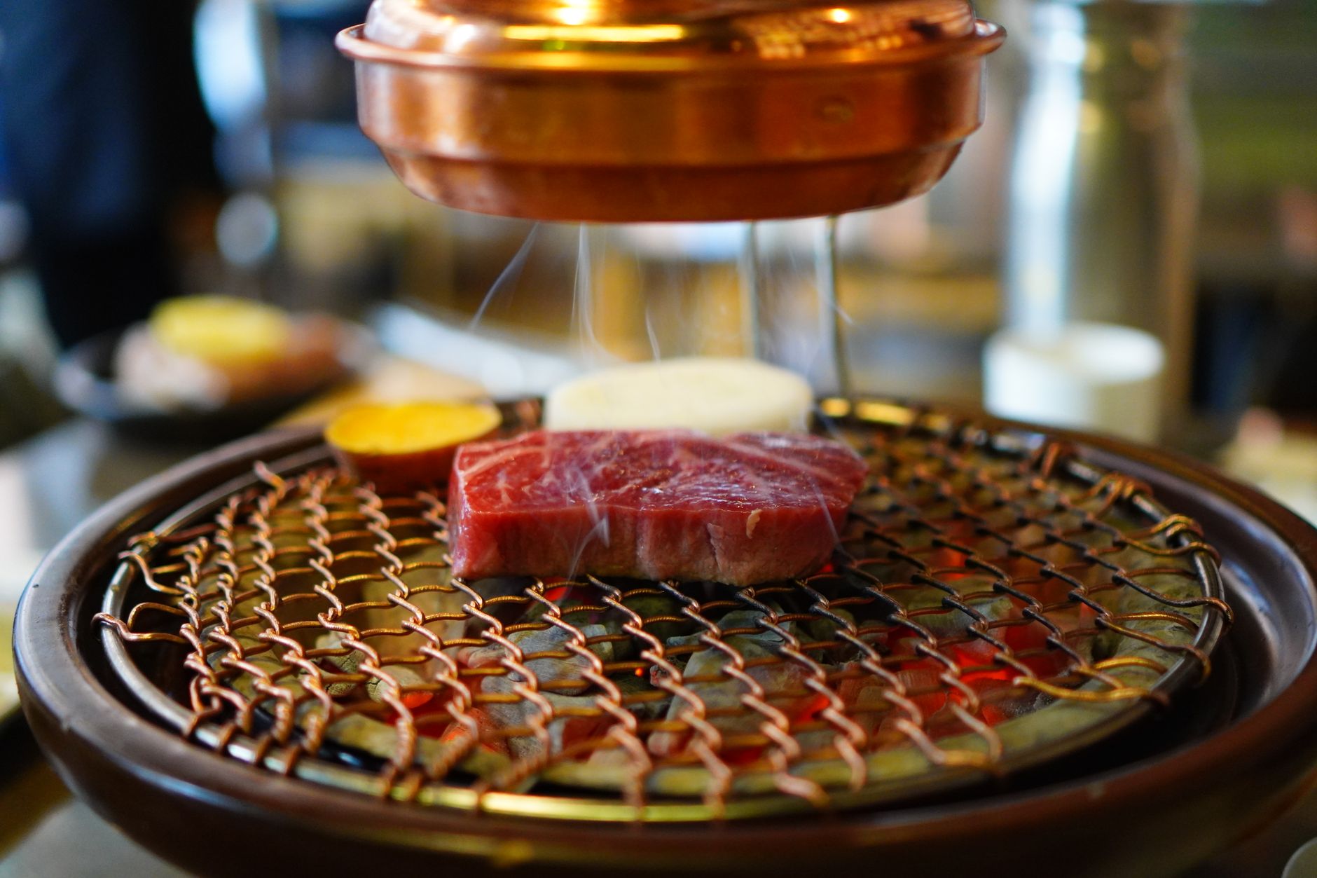 Close-up of raw beef on a Korean BBQ grill, sizzling under a copper hood in a restaurant setting.