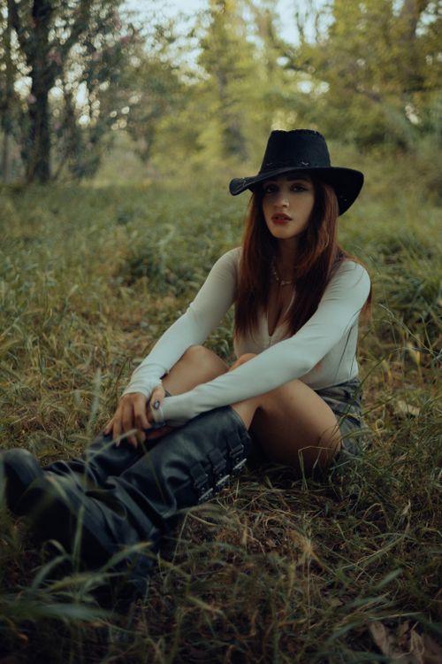 Free A fashionable woman in a black hat and boots sitting on grass in a natural setting. Stock Photo