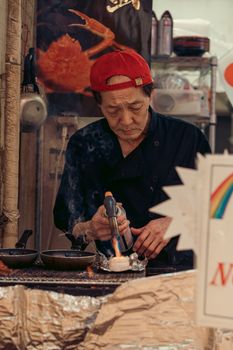 Elderly chef skillfully prepares food with a blowtorch at a street market, wearing a red cap.