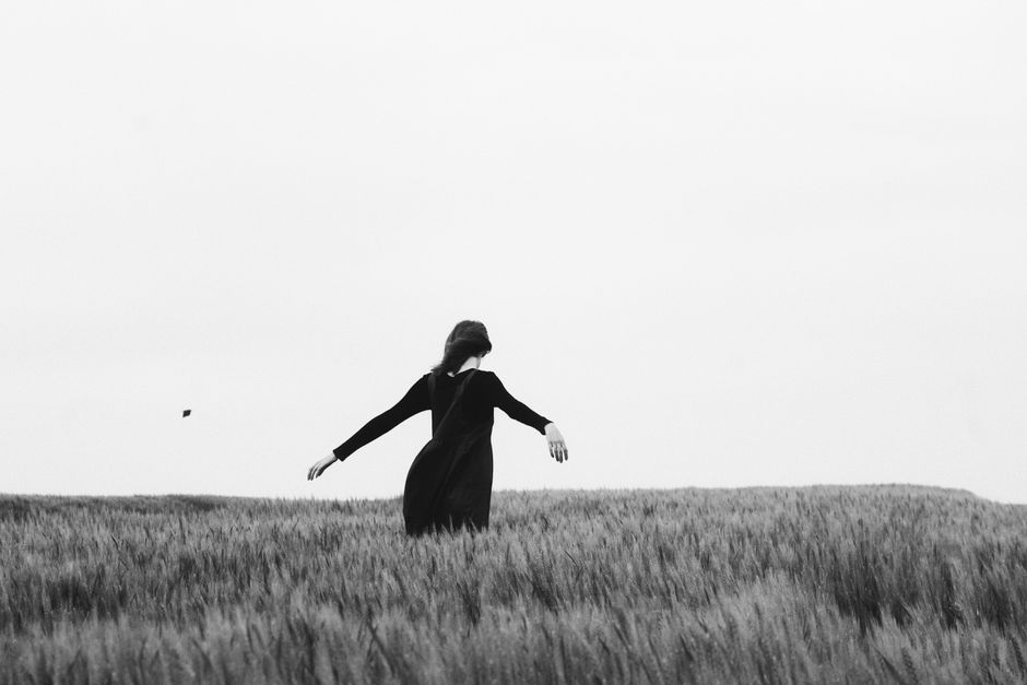 A woman in a black dress walks with arms outstretched in a serene wheat field.