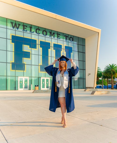 A woman in graduation gown and cap standing in front of a building with a welcome to fiu sign