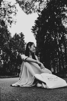 一张黑白照片中，一名身着连衣裙的女子坐在林间小路上，四周树木环绕