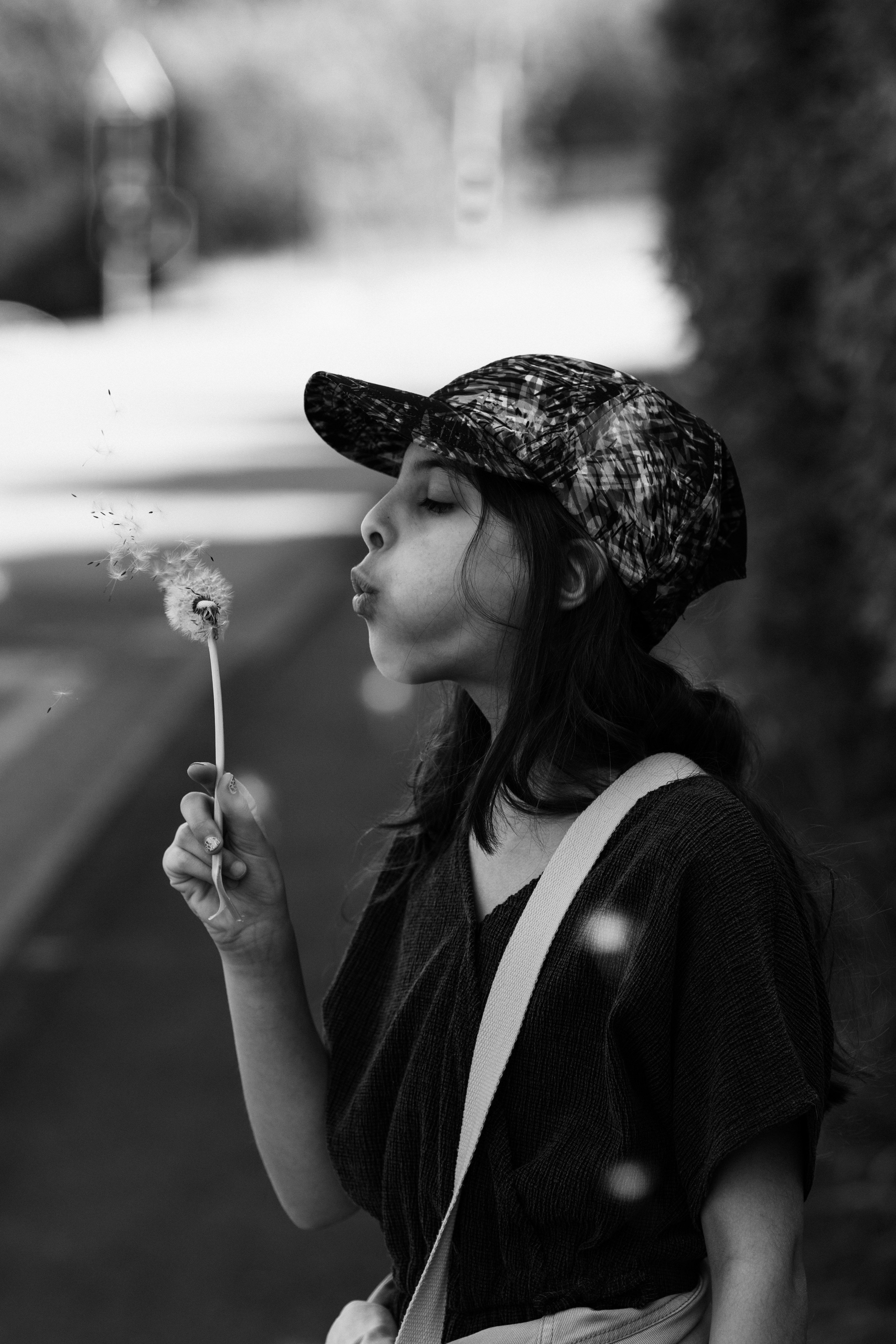 A young girl in a baseball cap blowing a dandelion in a serene outdoor setting.