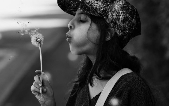 A young girl in a baseball cap blowing a dandelion in a serene outdoor setting.