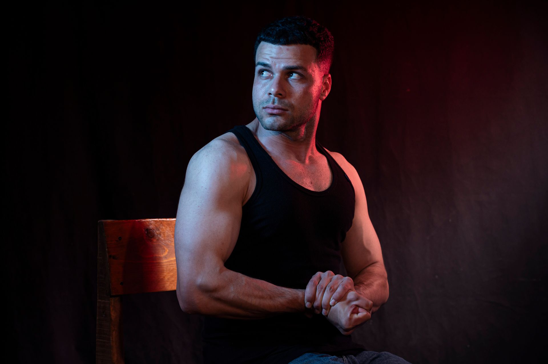 A muscular man in a black tank top poses against a dark background with dramatic lighting.