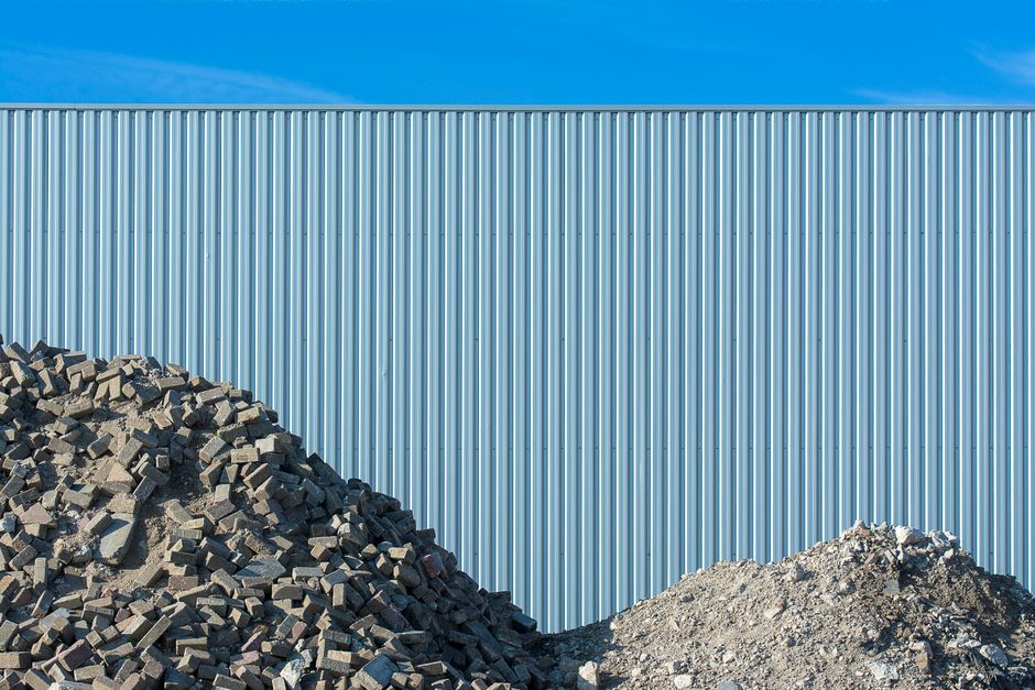 A pile of rubble in front of a large corrugated metal fence under a clear blue sky.