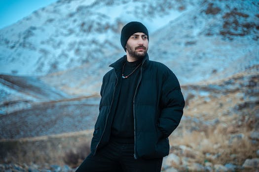 Man in black jacket and hat stands in snowy mountainous landscape during winter.
