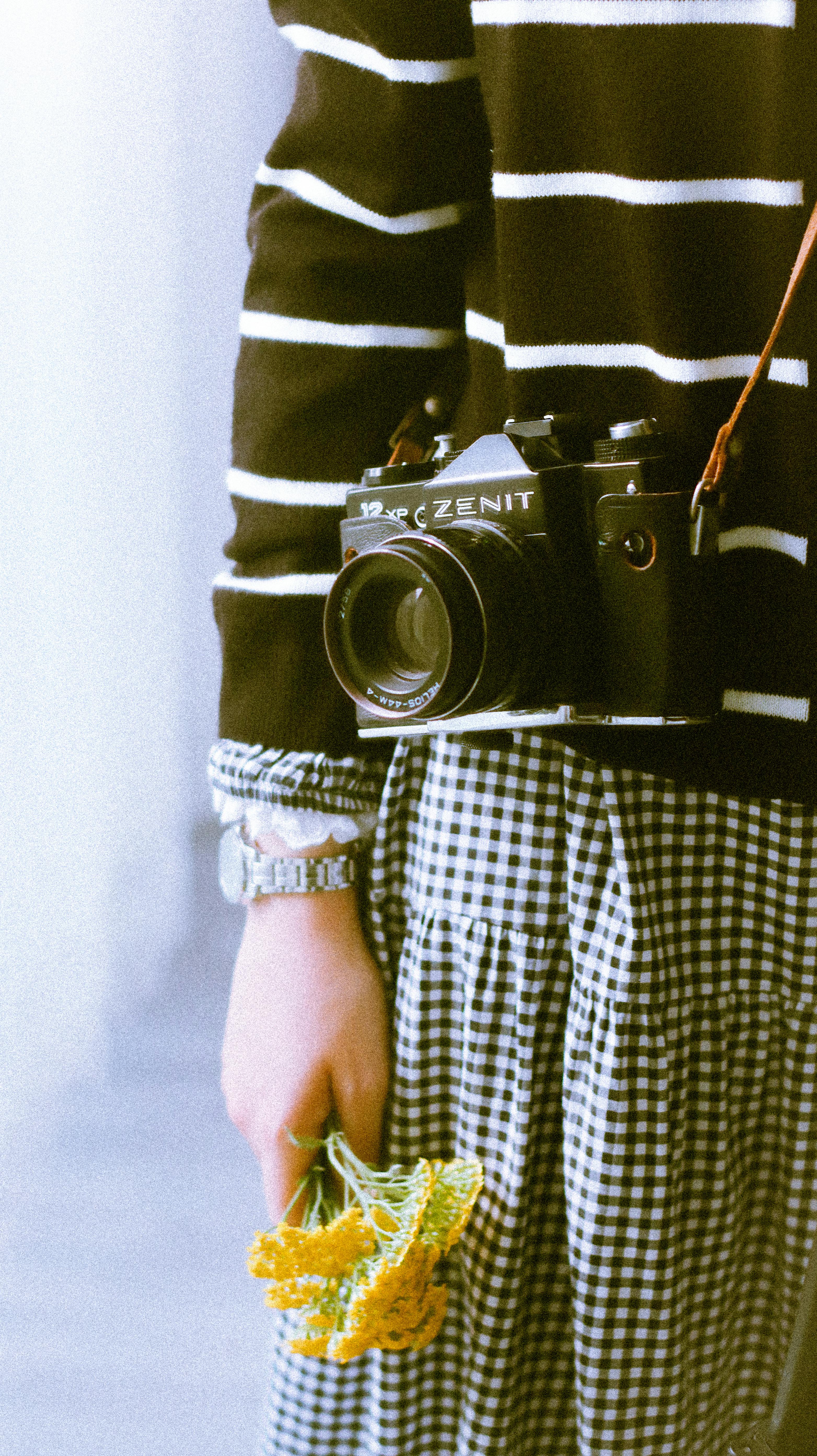 Woman standing indoors holding flowers and a vintage camera, wearing a striped sweater and gingham skirt.