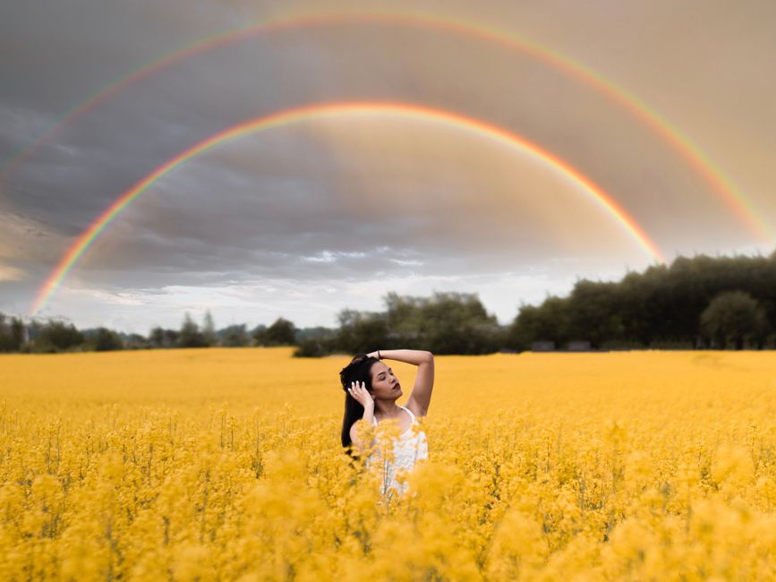 Serene woman posing in a vibrant mustard flower field with a stunning double rainbow overhead.