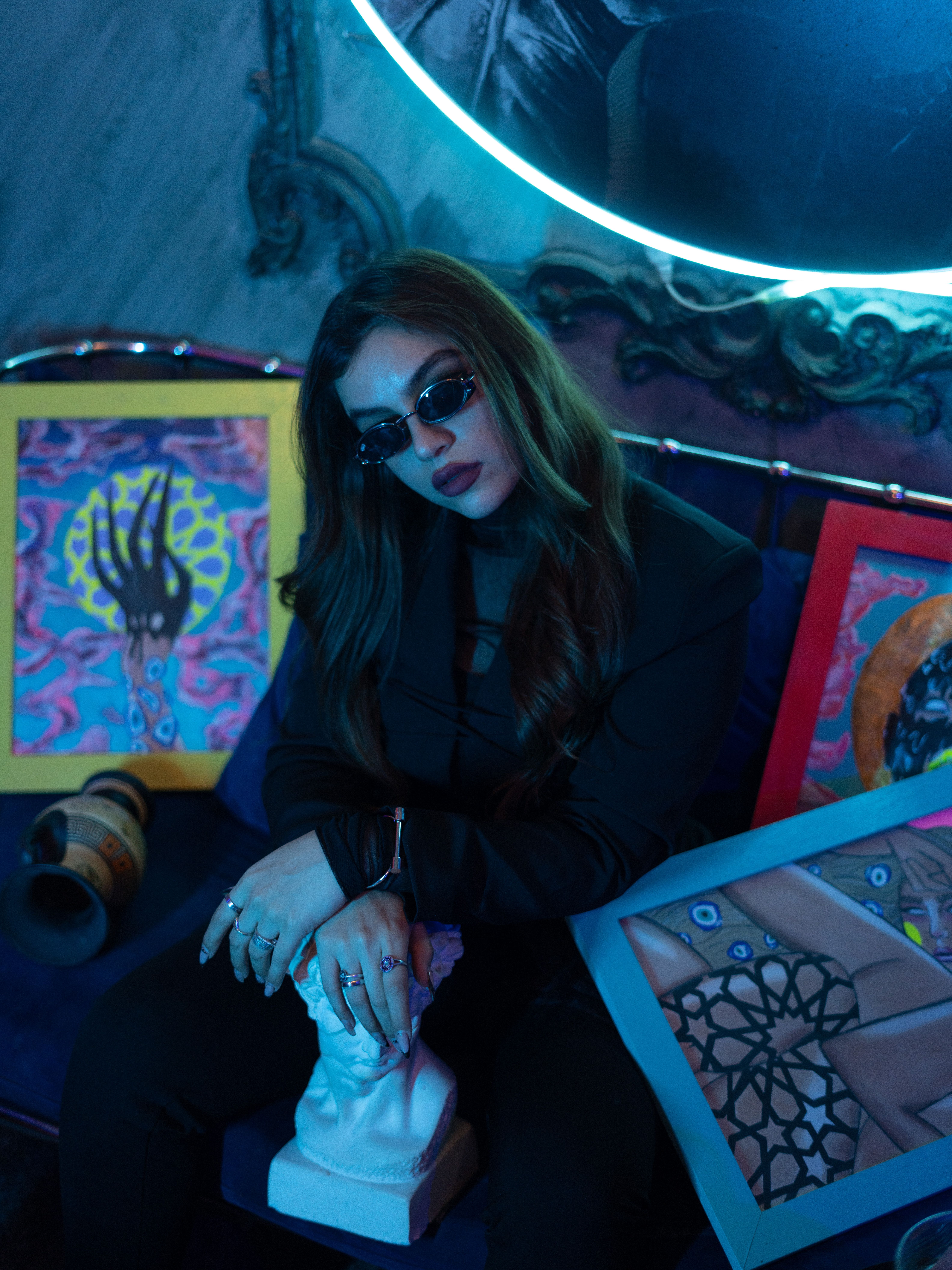 Chic woman in sunglasses posing with colorful art pieces in a dimly lit room.