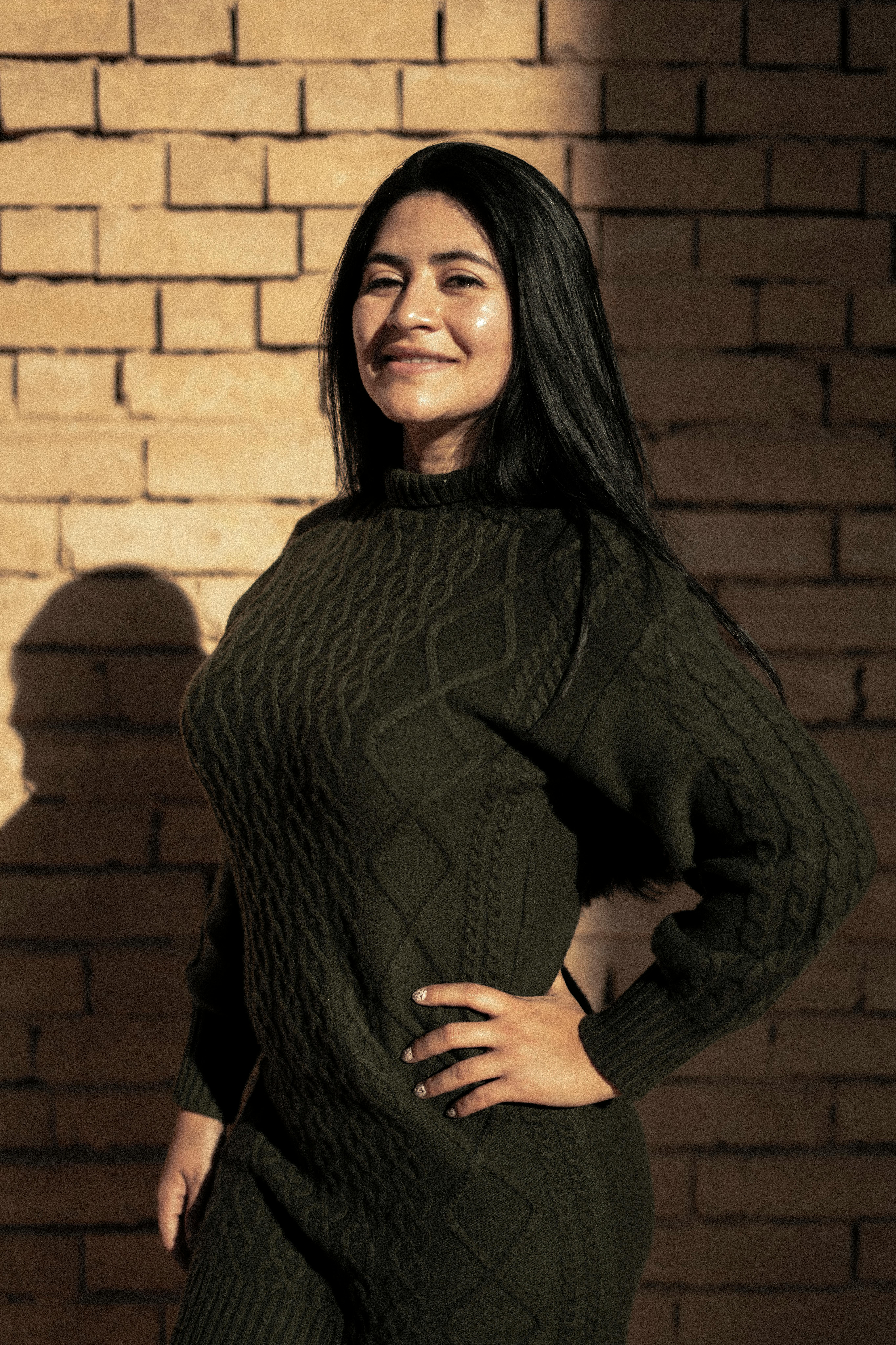 Free A cheerful woman in a textured sweater stands against a sunlit brick wall, exuding warmth and confidence. Stock Photo