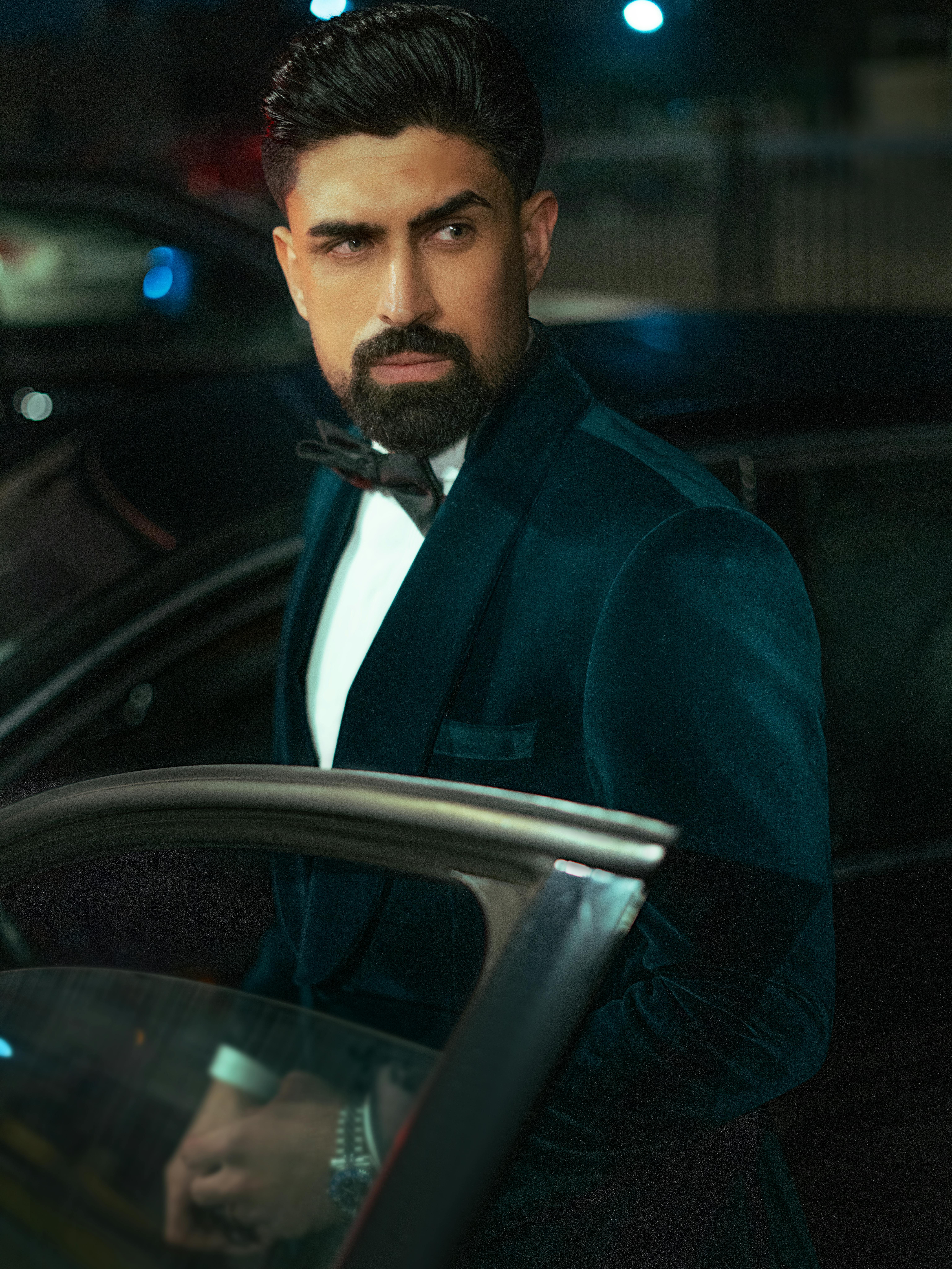 Stylish man in a tuxedo stands by a car, showcasing elegance and sophistication.