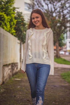 A young woman smiling and posing outdoors in jeans and a knitted sweater.