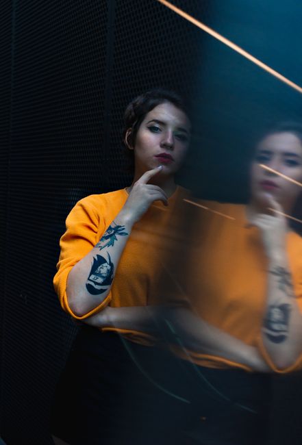 Artistic portrait of a woman in yellow sweater with tattoos, posing by a mirror for a reflective effect.