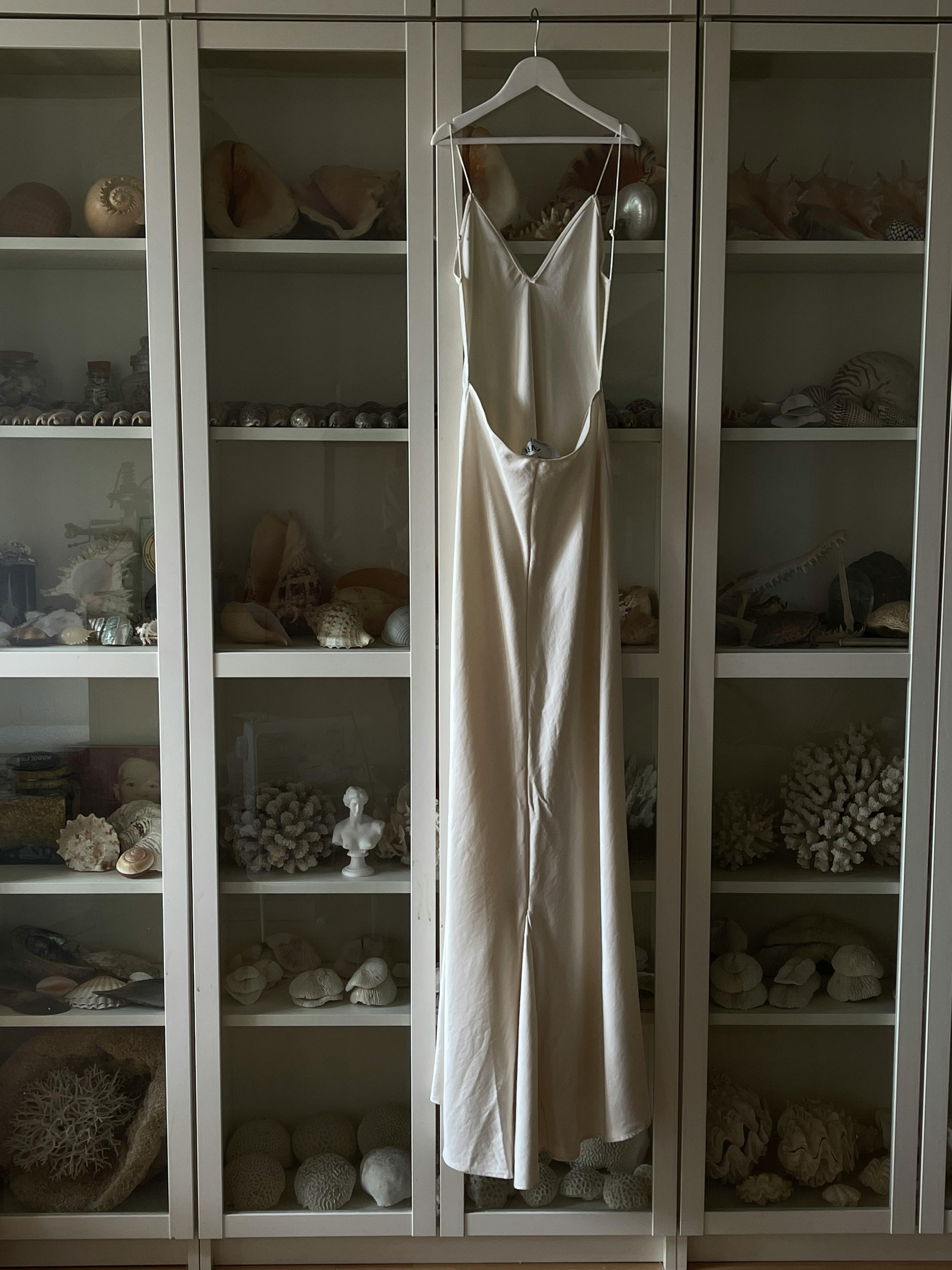 A minimalist white dress on a hanger against a seashell-filled cabinet, evoking coastal elegance.