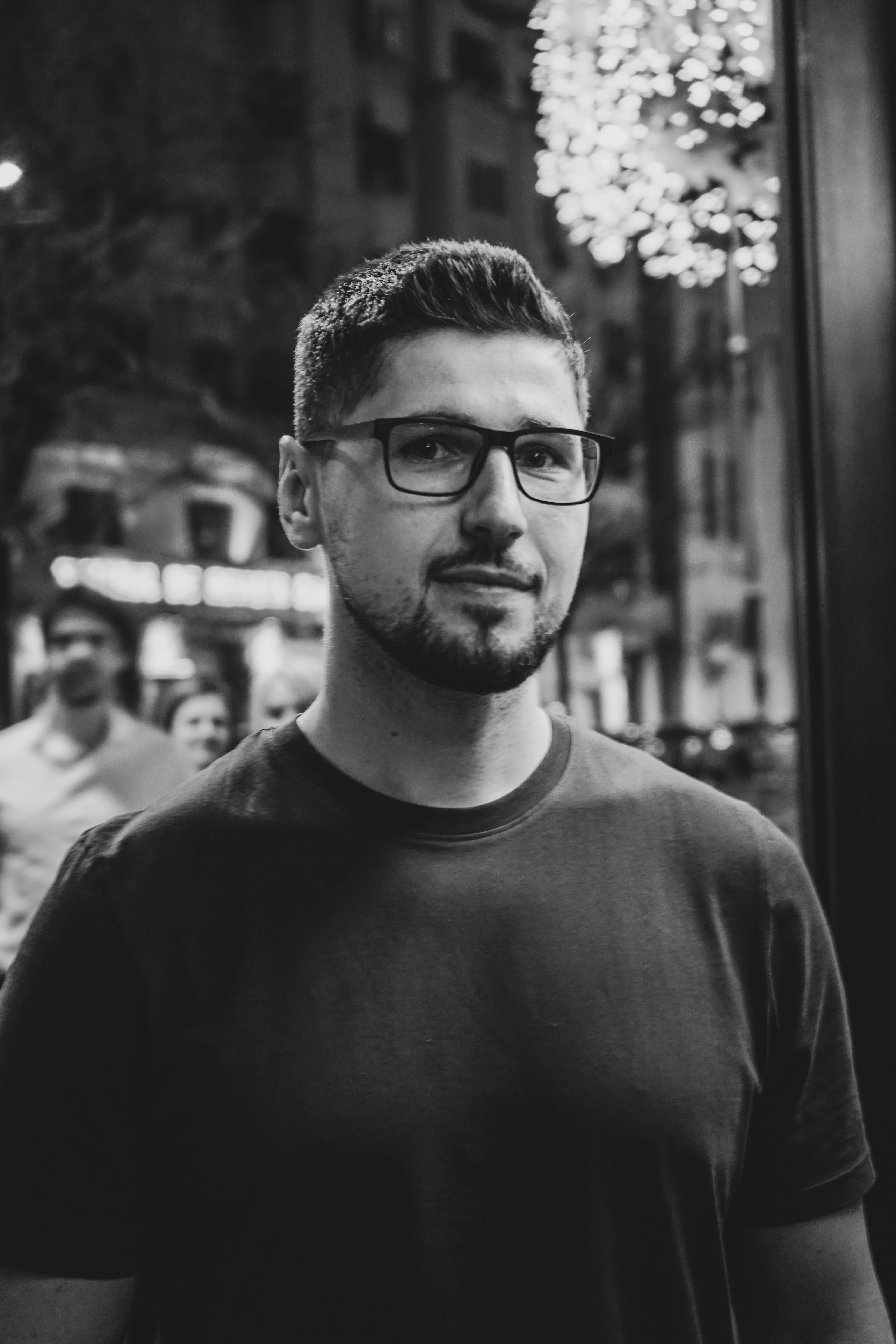 A thoughtful man wearing glasses in a night street scene, captured in black and white.