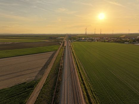 Railway track running through lush fields and wind turbines under a golden sunset sky.