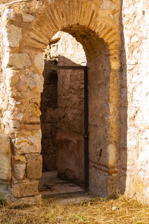 Free Rustic stone archway with open iron gate leading to ancient ruins, bathed in warm sunlight. Stock Photo