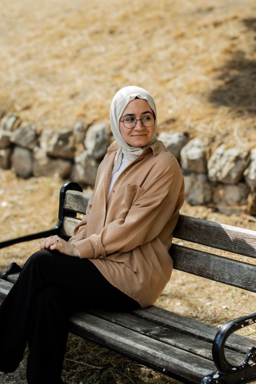 Free A woman wearing a hijab and glasses sits on a wooden park bench, enjoying a sunny day outdoors. Stock Photo