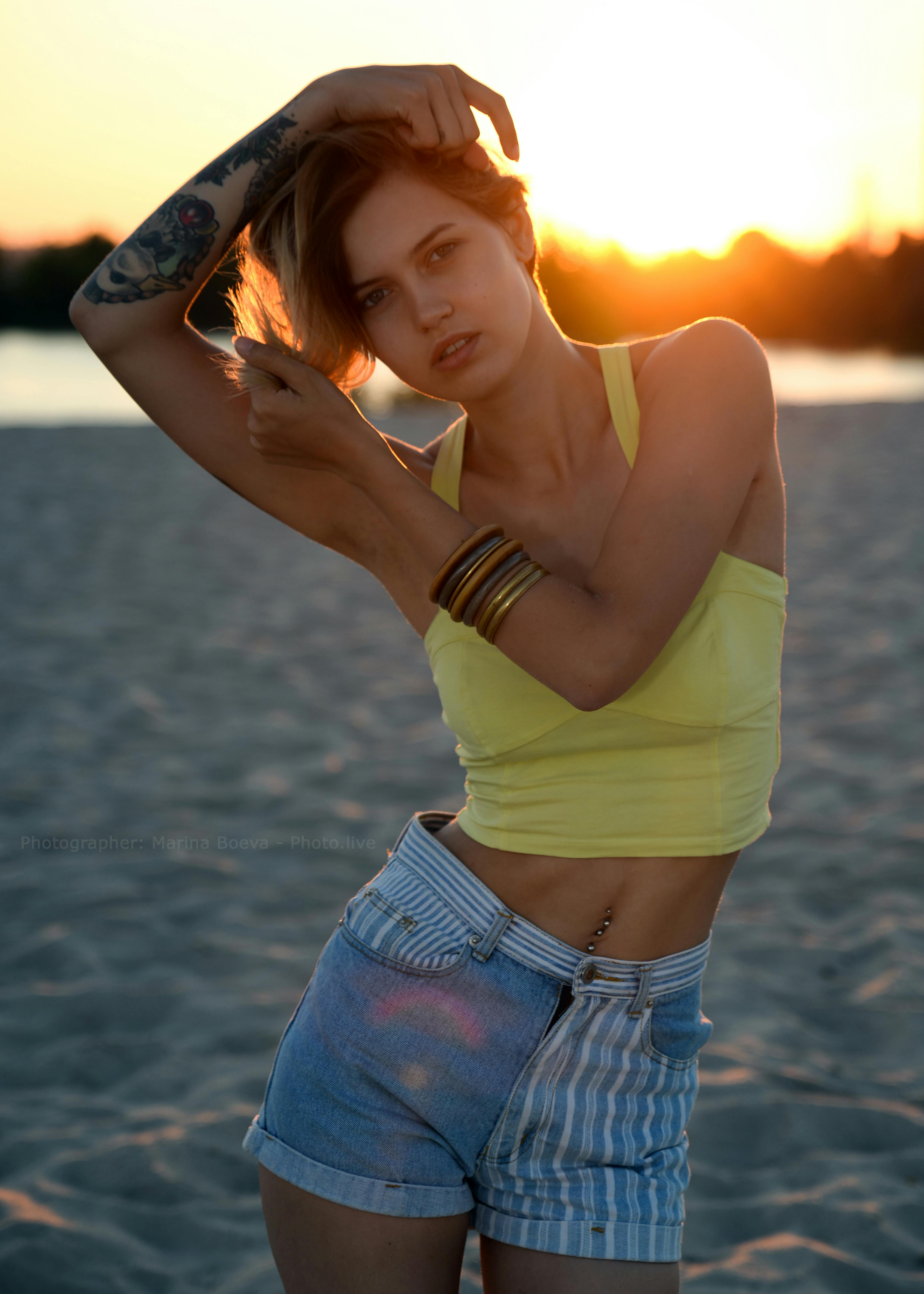 A young woman with a tattoo gracefully poses on a sandy beach at sunset, showcasing trendy summer fashion.