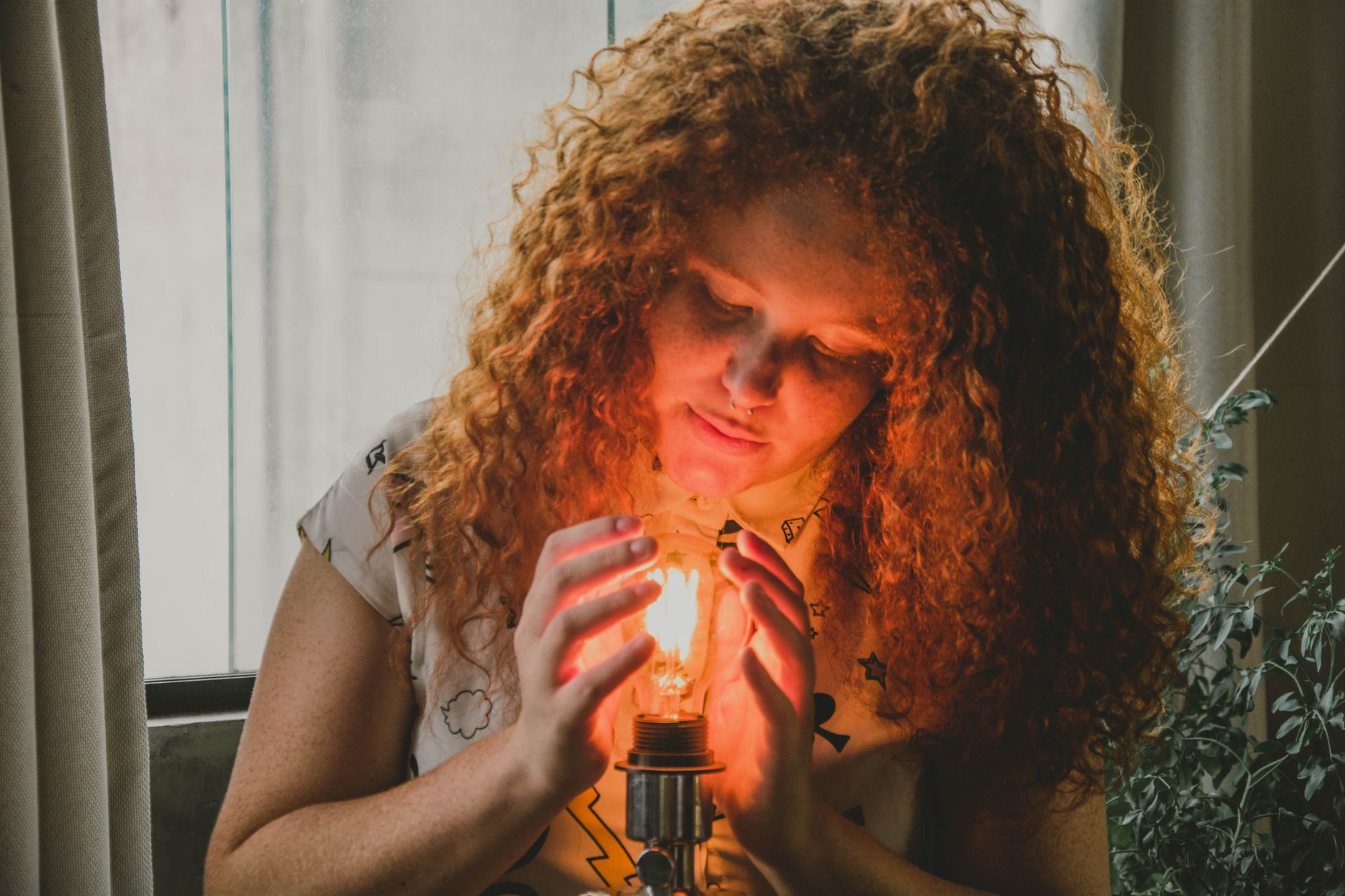 A woman with curly hair contemplates a glowing light bulb indoors.