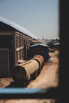 A rustic railway freight yard featuring tank cars next to a warehouse, emphasizing industrial logistics.