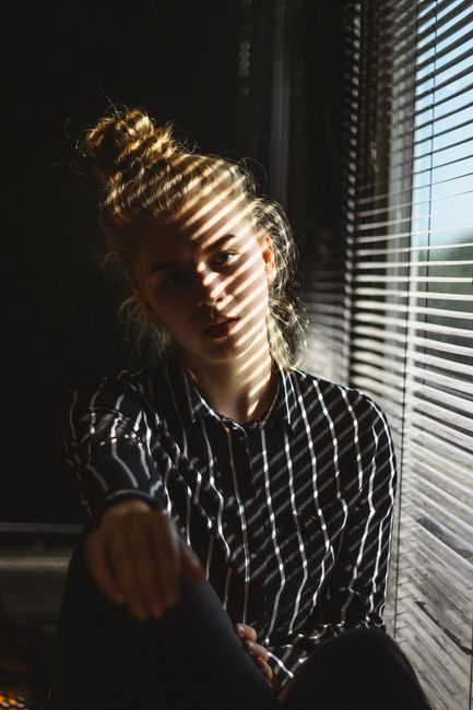 A contemplative woman sits by window blinds casting shadows inside.