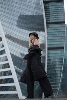 Fashionable woman in coat and hat posing against modern city architecture.
