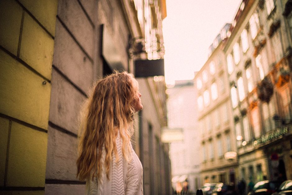A woman with long blonde hair looks up at city buildings, enjoying the urban atmosphere on a sunny day.