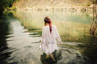 一位身着白色连衣裙的女子，涉入被大自然环抱的平静湖泊，营造出宁静而引人沉思的景象