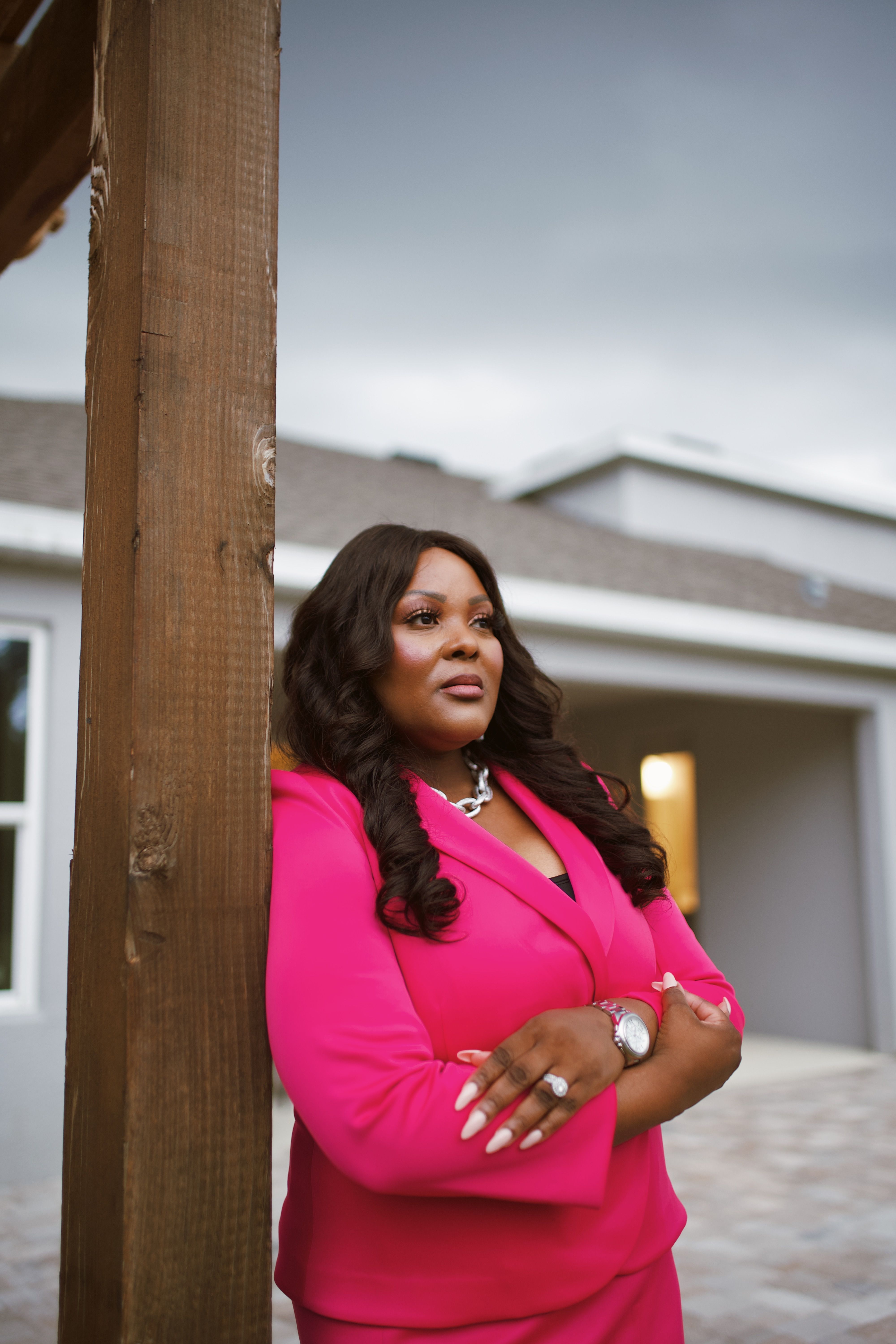 Confident woman in a stylish pink suit posing elegantly outdoors by a wooden pillar.