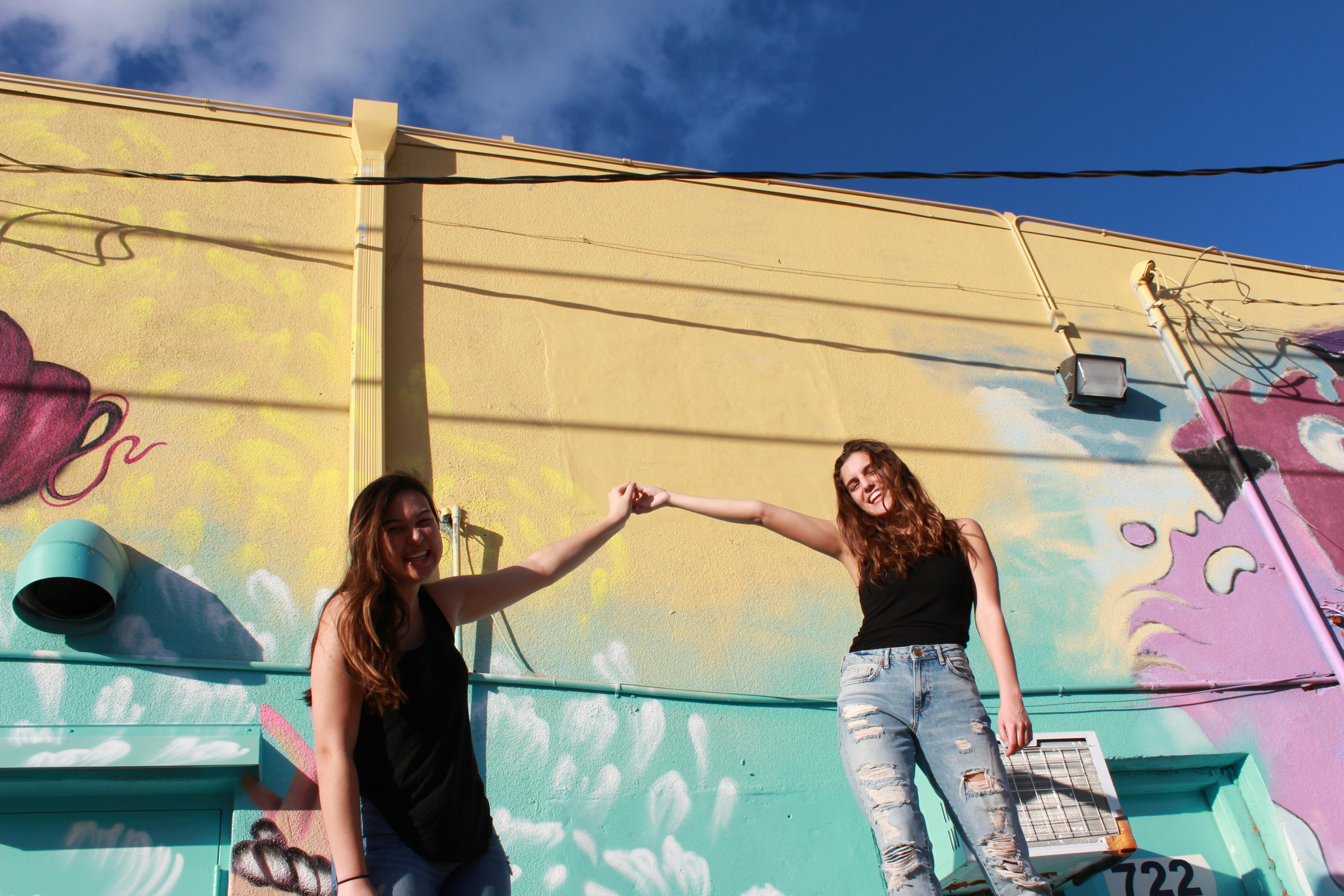 photo of two laughing women holding hand while standing near graffiti wall