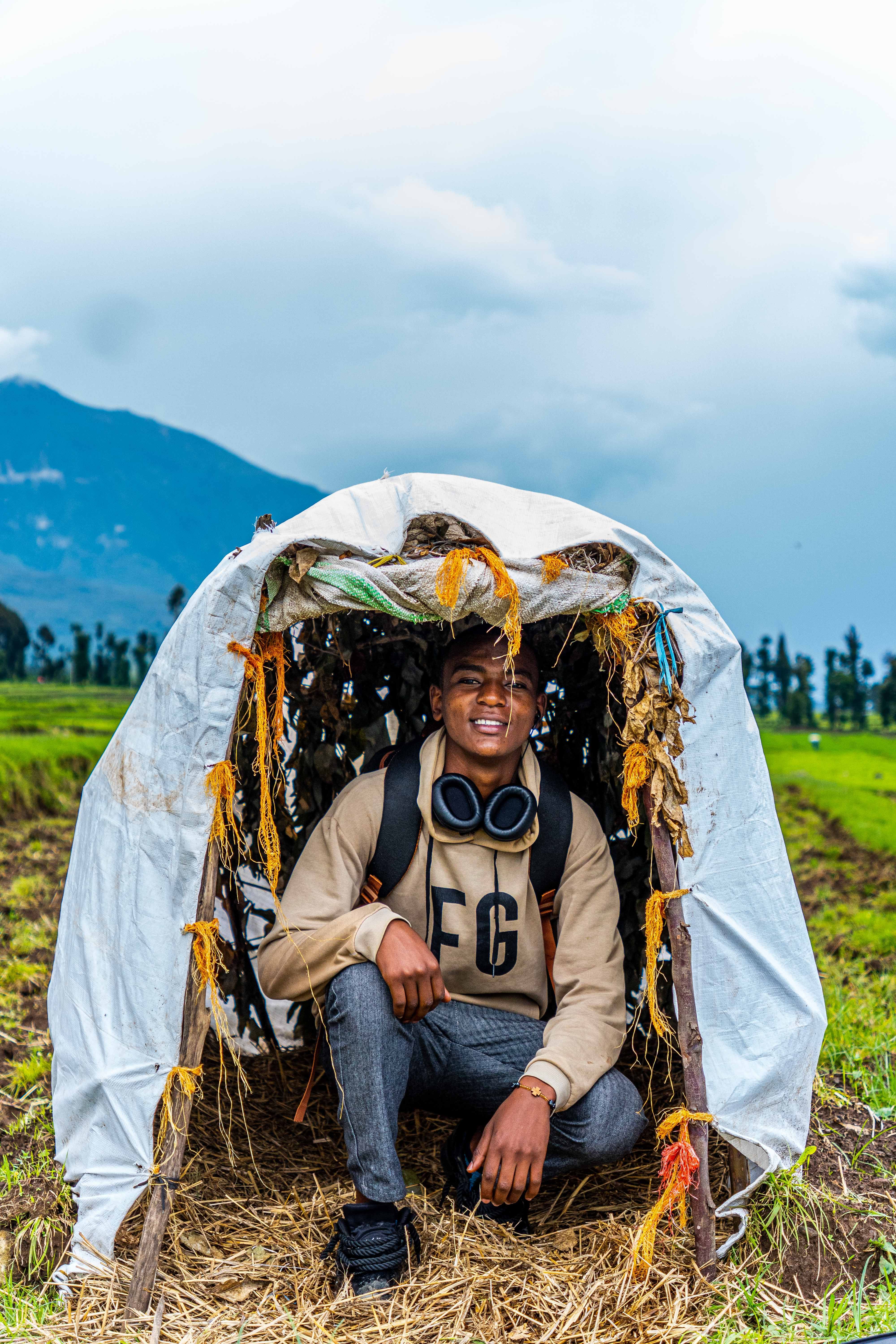 A young man enjoying outdoor adventure crouching in a makeshift tent with scenic mountain backdrop.