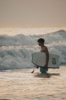 A young man holding a bodyboard, standing in the ocean waves during sunset, ready to surf.