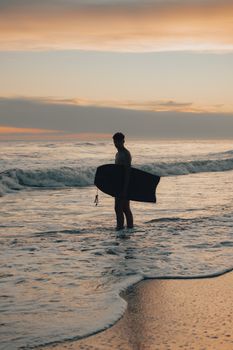 A surfer stands with a surfboard at sunset on a peaceful beach, capturing the essence of leisure and adventure.