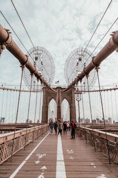 People walking on the iconic Brooklyn Bridge in New York City with a cloudy sky above.