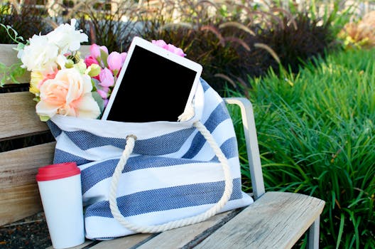A tablet in a striped tote bag with flowers on a park bench, ideal modern outdoor scene.