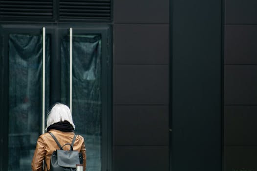 A woman with white hair stands outdoors facing a modern building's glass door.