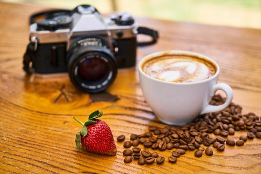A cup of cappuccino with coffee beans, a camera, and a strawberry on a wooden table.