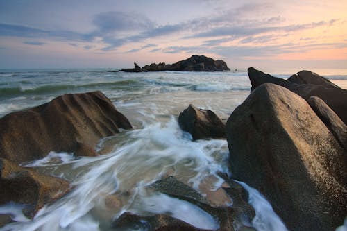 Sea Waves Crashing on Brown Boulders during Golden Hour Time Lapse Photo