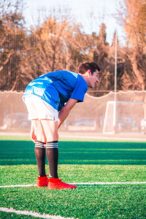 Free Male soccer player in blue jersey resting on a green field under sunlight. Stock Photo
