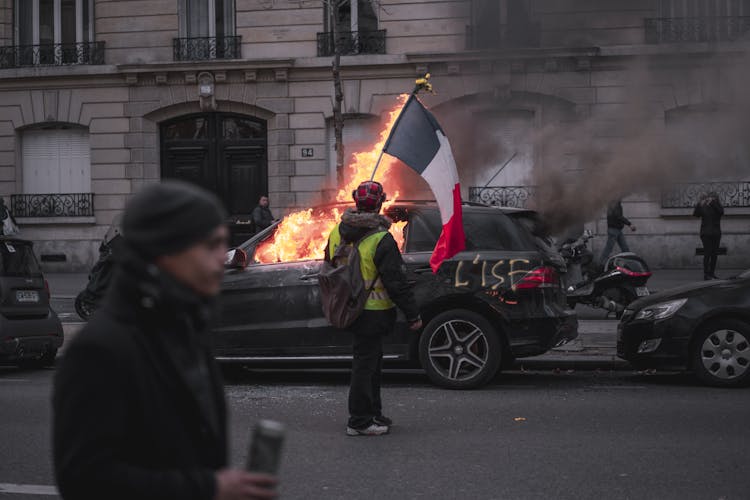 Man Holding A Flag Near A Burning Black Car