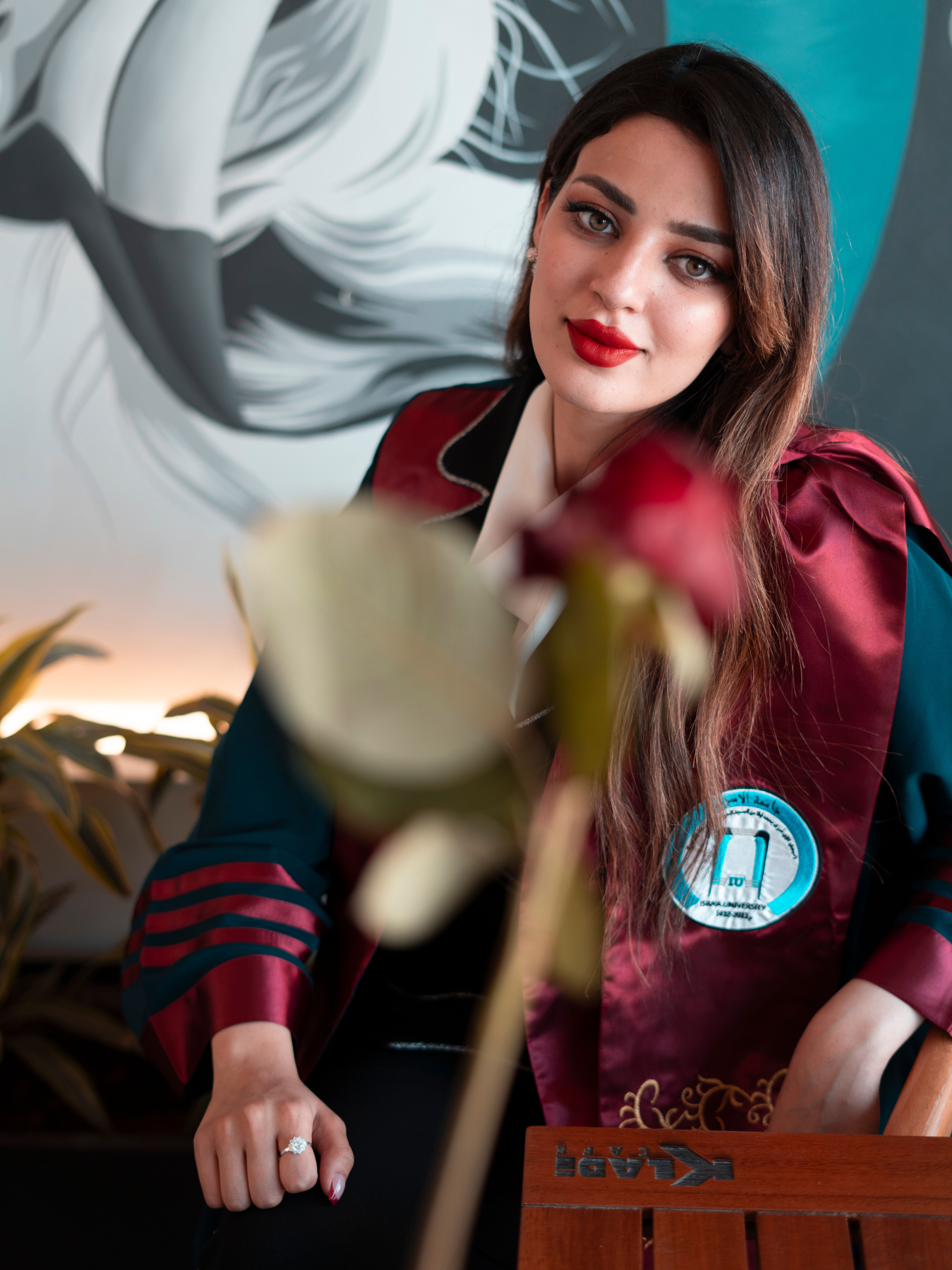 A young woman with red lips poses elegantly in graduation attire. Indoor setting with a rose in foreground.
