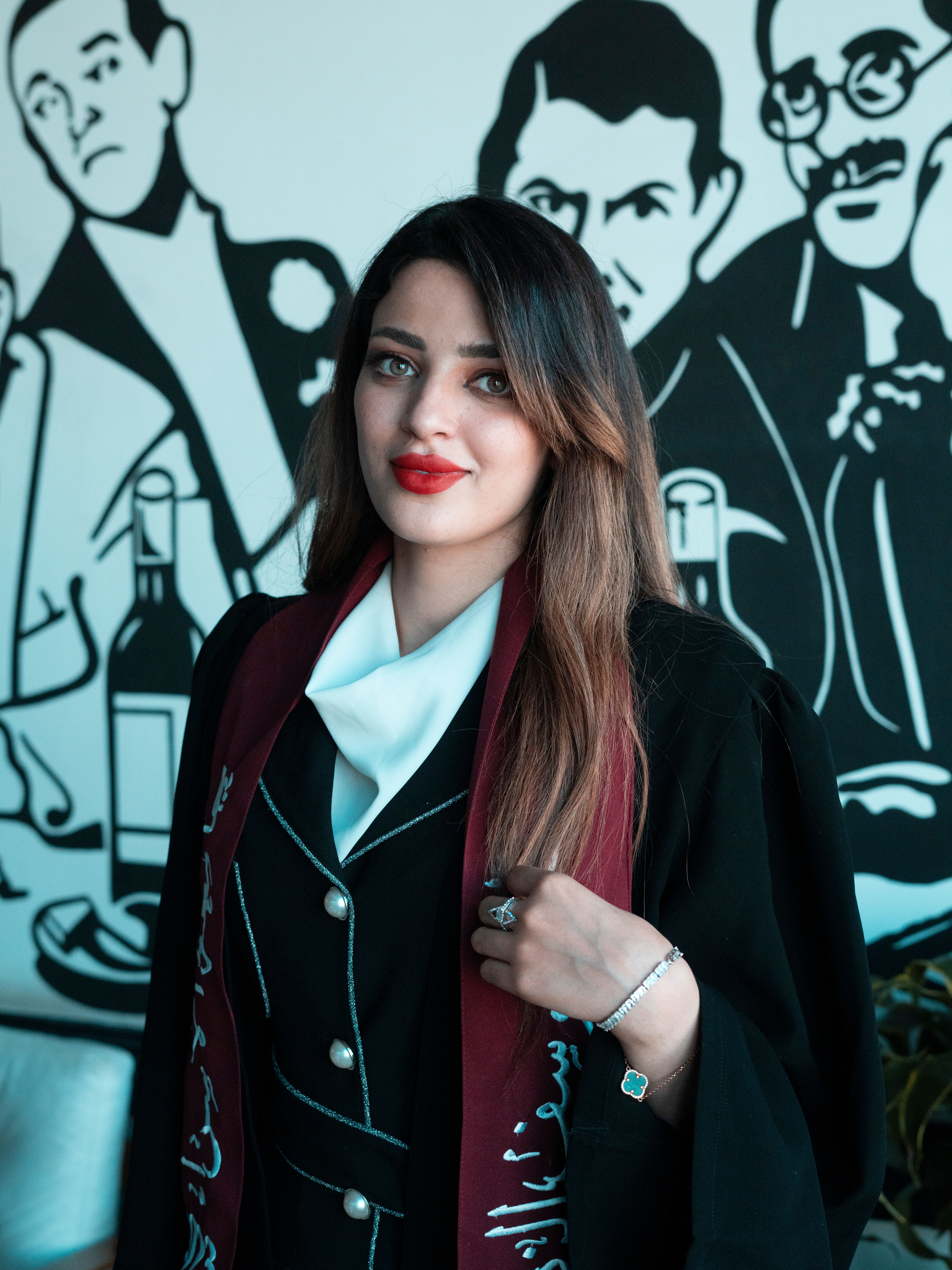 Young woman in graduation gown posing elegantly in a Baghdad cafe with artistic background.
