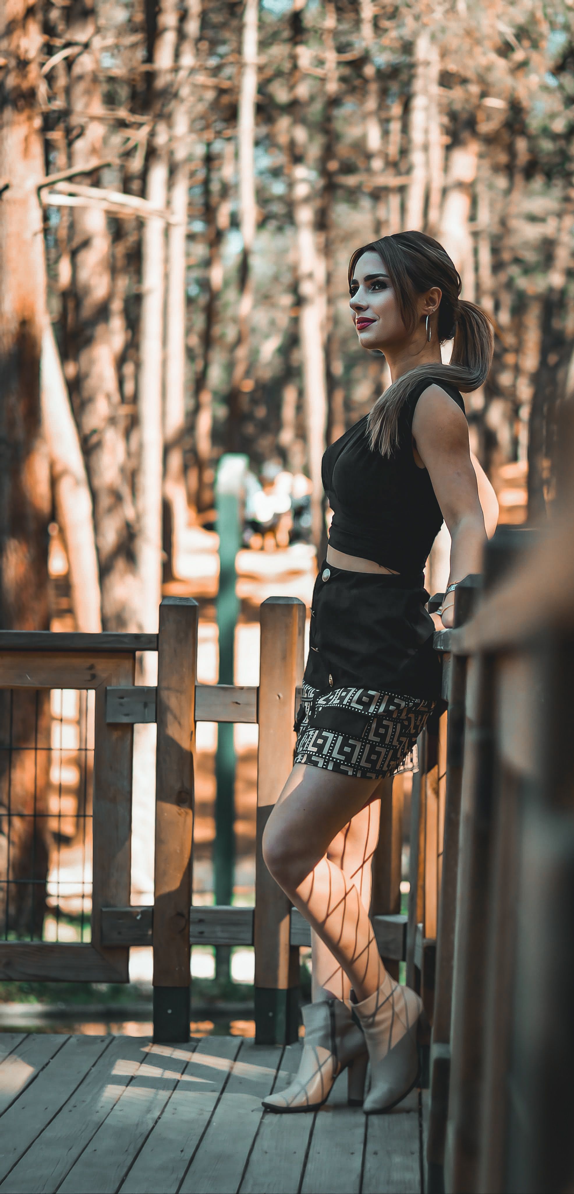 side view of woman wearing black crop top and skirt leaning against wooden railing in forest