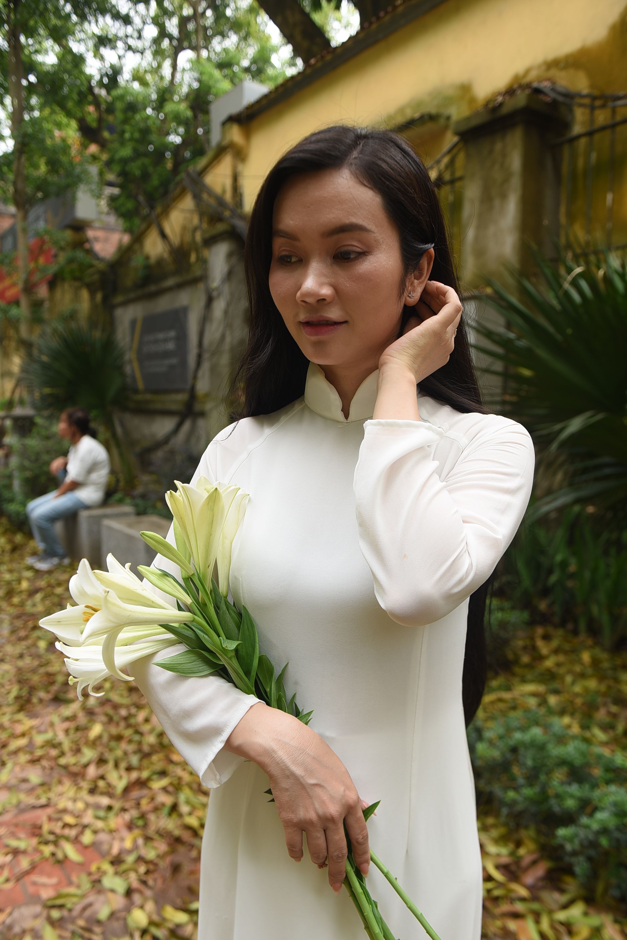 Asian woman in a white traditional dress holding lilies, in a serene outdoor setting.